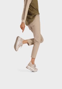 Scarpe da ginnastica beige con una parte superiore testurizzata, dettagli con lacci e una suola spessa bianca, abbinate a pantaloni tapered beige chiaro e un top verde oliva.