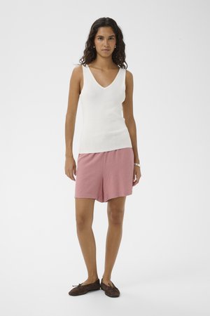 Femme debout portant un haut blanc sans manches à col en V, un short rose, des chaussures plates marron et une montre blanche au poignet sur un fond uni.