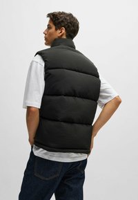 Black puffer vest with high collar and horizontal stitching, worn over a white t-shirt, paired with dark blue jeans. Smooth texture.