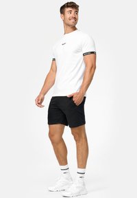 Weißes T-Shirt mit kurzen Ärmeln und schwarzen Akzentbändern an den Ärmeln, kombiniert mit schwarzen Shorts und weißen Sneakers mit gemusterten Socken.
