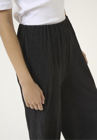 Black pleated pants made from lightweight fabric, featuring an elastic waistband and a textured surface, shown from the side with a hand on the waist.