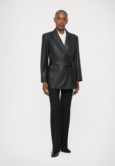 Čierny kožený blejzer s obalom, opaskom v páse a dvoma prednými vreckami. Nosený cez bielu košeľu a kombinovaný s čiernymi nohavicami.