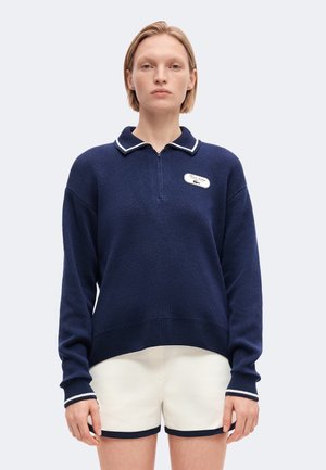 Femme portant un pull polo bleu marine à manches longues avec des bordures blanches et un short blanc avec des bords bleu marine, debout devant un fond clair.