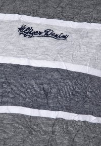 Pruhovaná látka v námořnické modré a bílé barvě s pomačkanou texturou; obsahuje vyšitý logo "Hilfiger Denim" v tmavě modré barvě na světlejším úseku.