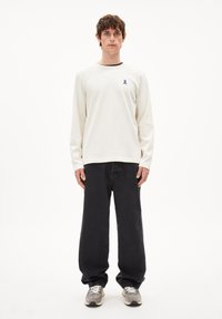 Witte lange mouwen shirt met een klein blauw lint op de borst, gecombineerd met zwarte wijde jeans en grijze sneakers. Simpel ontwerp en relaxte pasvorm.