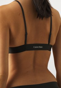 Svart bh med tynne stropper og en jevn tekstur. Har et bredt elastisk bånd med "Calvin Klein" logo bak. Minimalistisk design.