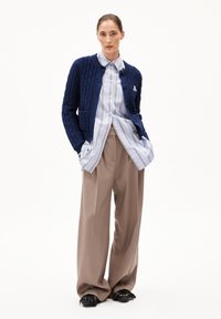 Dunkelblauer strukturierter Cardigan mit Rundhalsausschnitt, ausgestattet mit einer Vordertasche und Knopfverschluss; kombiniert mit einem hellblauen gestreiften Hemd und beigen Hosen.