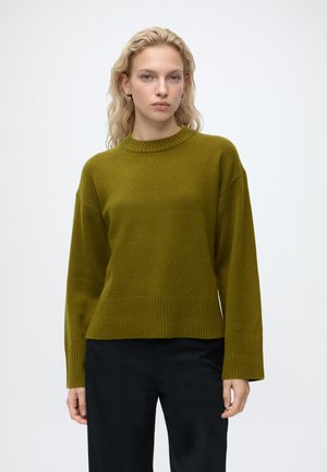 Femme blonde aux cheveux ondulés portant un pull en maille vert olive et un pantalon noir, debout devant un fond blanc uni.