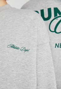 Grijze sweatshirt met een textuurfinish, met groen geborduurde tekst "Athletic Dept." op de mouw en "SUN" in grote letters op de achterkant.