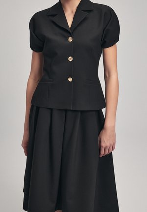 Woman wearing short-sleeve black blazer with gold buttons and matching black skirt, standing against a plain light gray background.