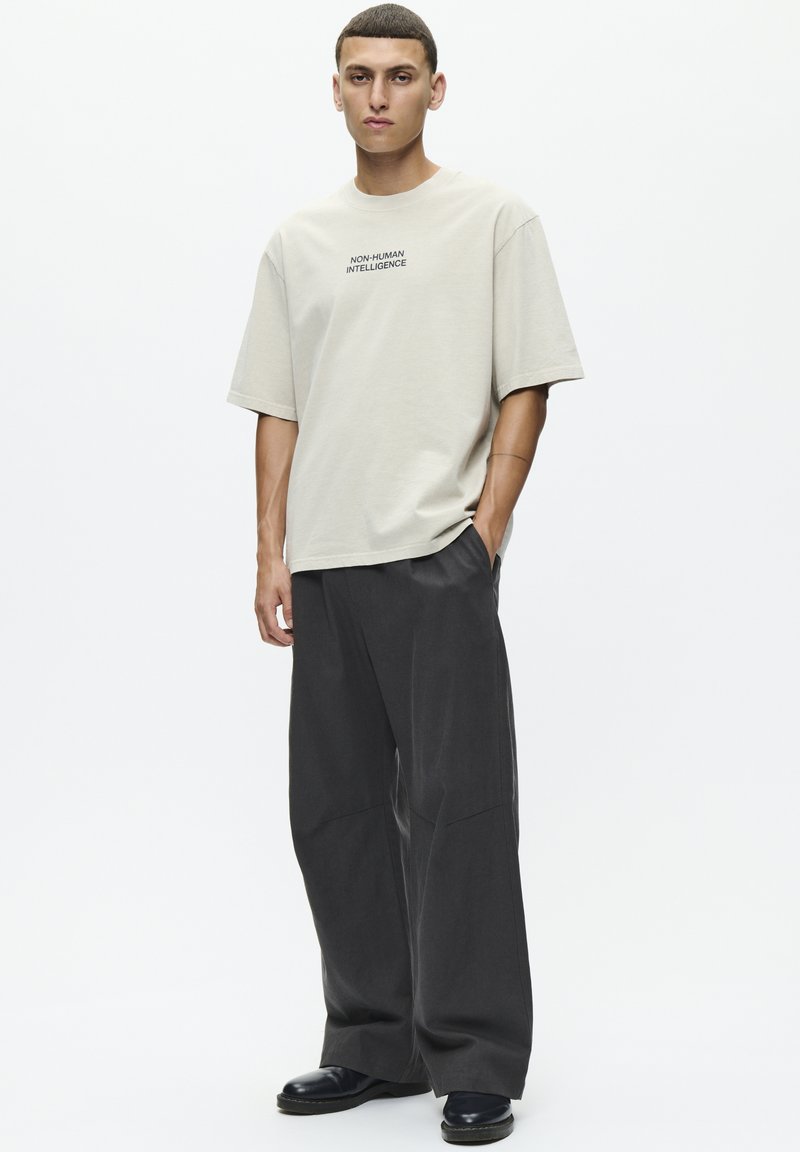 T-shirt beige clair avec le texte "INTELLIGENCE NON-HUMAINE", associé à un pantalon gris foncé ample et des chaussures noires. Coupe simple et décontractée.