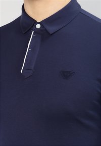 Marinblå polo skjorta i bomullsblandning, med klassisk krage, treknappstängning och en diskret logotyp på vänster sida.