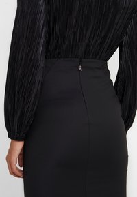 Svart plisserad topp med långa ärmar som är kombinerad med en figursydd svart kjol med en dold dragkedja och en liten metallisk dragring på baksidan.