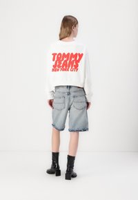 Bílá mikina se velkým červeným nápisem "TOMMY JEANS NEW YORK CITY." V kombinaci s vytvořenými lemy, světle modrými denimovými kraťasy a černými kotníkovými botami.