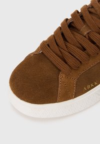 Brown suede sneaker with round toe, textured surface, and prominent laces. White rubber sole features geometric pattern and brand logo.
