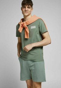 Jonge man die een groen Jack & Jones T-shirt en bijpassende korte broek draagt, met een oranje trui om zijn schouders geknoopt.
