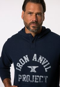Sudadera azul marino con un bolsillo frontal, con un escote redondeado y un estampado gráfico que dice "IRON ANVIL PROJECT" en texto blanco desgastado.