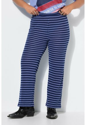 Personne portant un pantalon bleu à motifs et des bottines noires, debout contre un mur clair uni, les mains ajustant la taille.