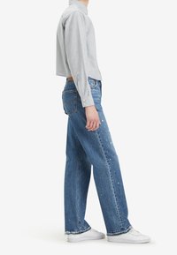 Camisa de rayas recortada con puños de botones combinada con unos vaqueros azules holgados que presentan un acabado desgastado y un dobladillo deshilachado, complementados con zapatillas blancas.