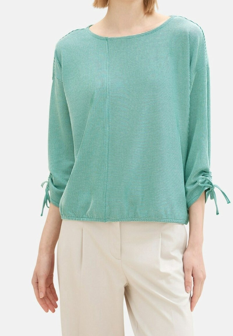 Žena na sobě má dlouhý rukáv zeleno-bílého pruhovaného topu s nařasenými detaily a světle béžovými kalhotami.