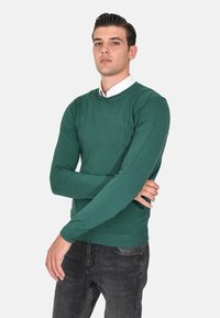 Maglione verde a maniche lunghe in morbido tessuto knit, colletto a giro, e polsini a costine. Abbinato a jeans scuri, con una vestibilità slim.