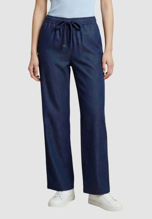 Donna che indossa pantaloni con coulisse blu scuro, top senza maniche azzurro chiaro e sneakers bianche, in piedi contro uno sfondo semplice.