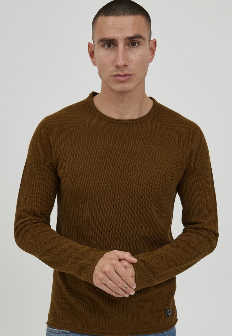 Brown knit long-sleeve shirt with a round neckline and textured fabric, featuring raglan sleeves and a small logo tag on the bottom left.