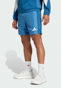 Los pantalones cortos deportivos azules cuentan con una cinturilla elástica, tres rayas blancas y un logo de Adidas bordado en blanco. Combinados con zapatos y calcetines blancos.