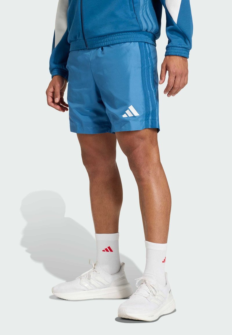 Los pantalones cortos deportivos azules cuentan con una cinturilla elástica, tres rayas blancas y un logo de Adidas bordado en blanco. Combinados con zapatos y calcetines blancos.