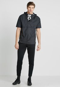 Schwarze Windjacke mit Kapuze und kurzen Ärmeln, ausgestattet mit einem halben Reißverschluss vorne und großem weißem Logo; kombiniert mit schwarzen, taillierten Hosen und Stollenschuhen.