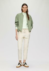 Veste bomber vert clair sur un pull à rayures blanches et vertes, assortie à un pantalon beige fuselé et des baskets à semelles épaisses bicolores.