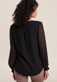 Blouse noire texturée avec de longues manches transparentes, des poignets froncés et un ourlet arrondi, présentant un empiècement lisse au dos.
