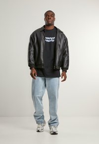 Black leather bomber jacket over a black t-shirt with white text, light blue jeans, and silver athletic shoes with black accents.