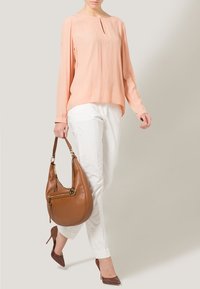 Femme portant un chemisier à manches longues couleur pêche, un pantalon blanc, des talons hauts marron, tenant un grand sac à main en cuir marron à ses côtés.