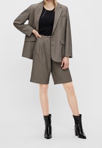 Veste à carreaux marron avec deux boutons, poches inclinées et short marron plissé. Complété par des bottines noires.