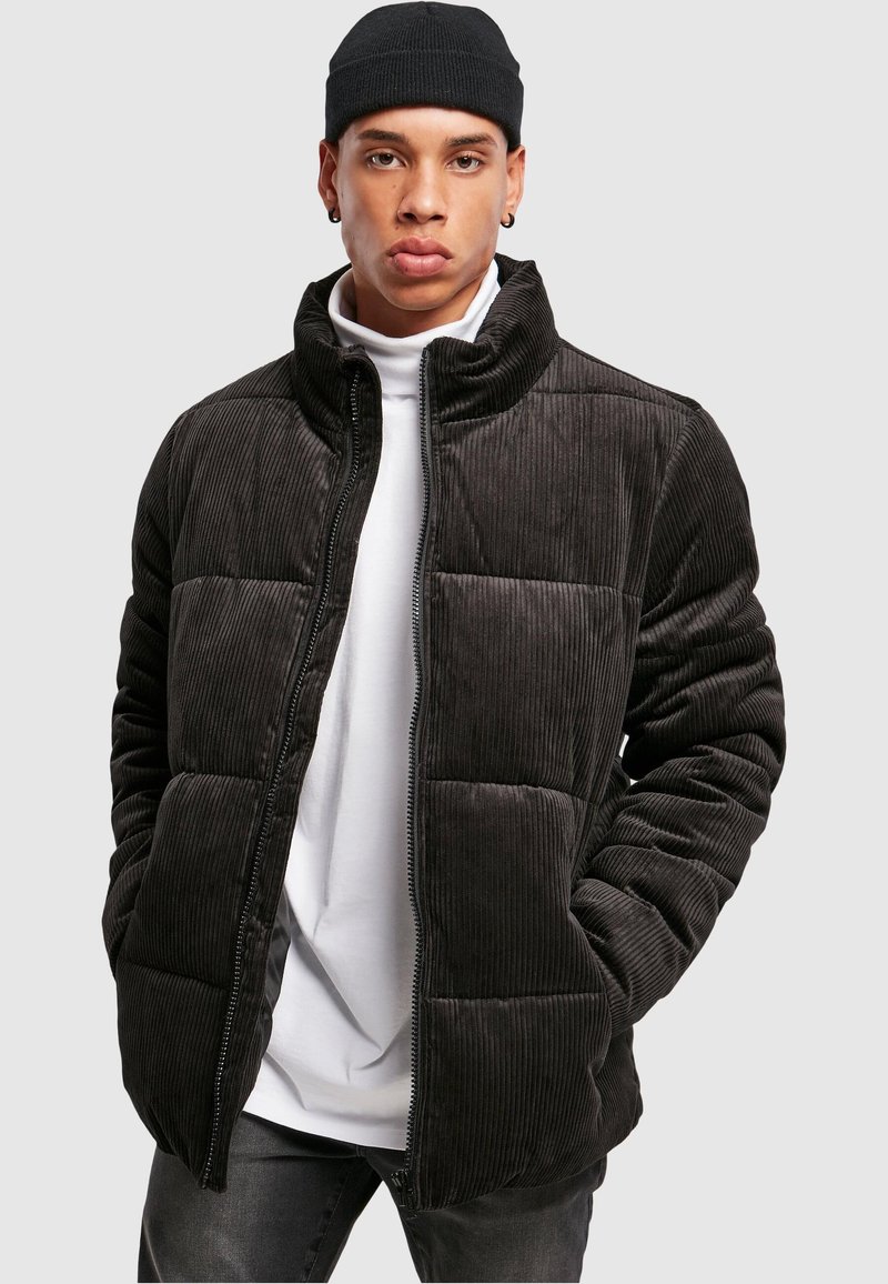 Abrigo tipo puffer de pana en gris oscuro con diseño acolchado, cuello alto y cierre de cremallera, combinado con un cuello alto blanco debajo.
