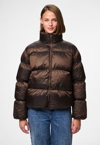 Brun puffjacka med hög krage, texturerat tyg, ribbade muddar och en central dragkedja, kombinerad med blå denimjeans.