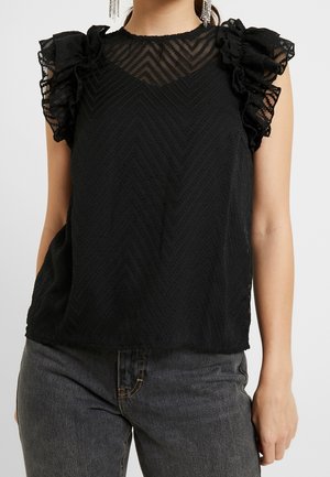 Femme portant un top noir sans manches en tissu transparent à motifs avec des épaules à volants, associé à un jean gris foncé.