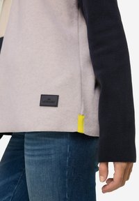 Giacca in felpa di colore grigio chiaro e blu navy con una finitura texturizzata, caratterizzata da un'etichetta nera e un'etichetta accentata gialla all'orlo.