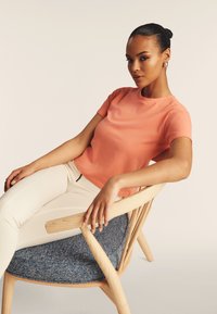 Camiseta de manga corta en color coral de tejido suave, combinada con pantalones beige de corte ajustado. Modelo sentado en una silla de madera con tapicería azul.