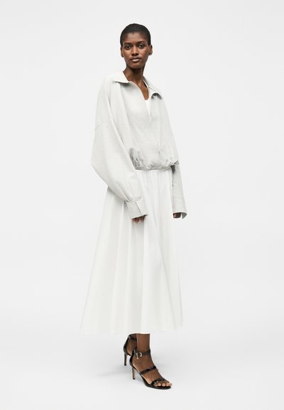 Γυναίκα στέκεται φορόντας ένα φαρδύ ανοιχτόγκρι σακάκι με μακριά μανίκια, μια λευκή ρέουσα midi φούστα και μαύρα σανδάλια με λουράκια και ψηλά τακούνια.