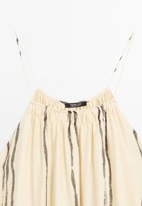 Débardeur couleur crème avec fines bretelles spaghetti, encolure élastique froncée et rayures noires verticales pour la texture. Étiquette de la marque visible.