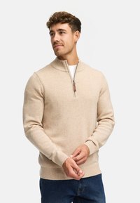 Beige stickad tröja med hög krage och halv dragkedja. Har ribbade ärmar och nederdel samt en mjuk textur. Lämplig för avslappnad klädsel.