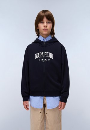 Marinblå huvtröja med vit text "NAPA PIJRI", frontdragkedja och fast capuchong. Buren över en ljusblå skjorta med krage och beige byxor.