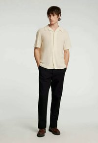 Rövid ujjú, krémszínű kötött póló gallérral, fekete nadrággal és barna cipővel párosítva. Egyszerű dizájn, finom textúrával.