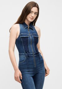 Νεαρή γυναίκα που φοράει αμάνικη εφαρμοστή ολόσωμη φόρμα από τζιν με φερμουάρ μπροστά, γιακά και δέσιμο κορδονιών στο στήθος.