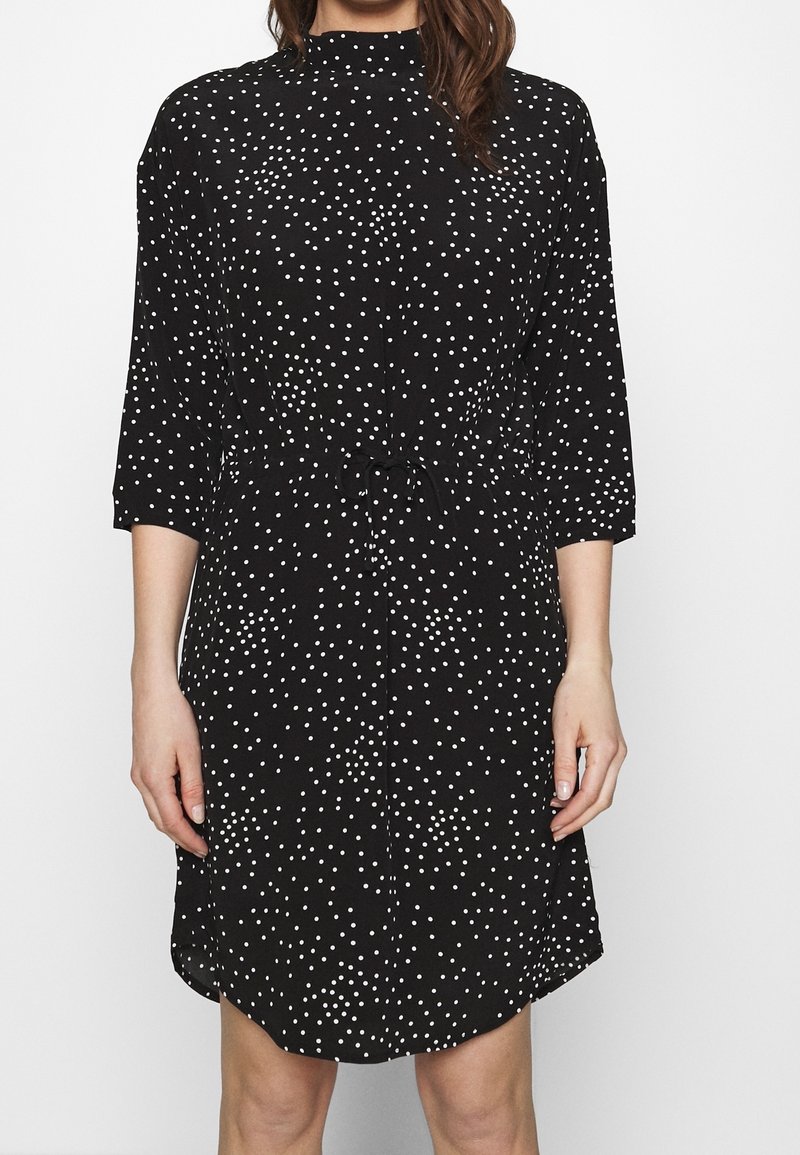 Femme portant une robe noire longueur genou à pois blancs, manches trois-quarts, avec un détail de taille noué, montrée du cou aux genoux.