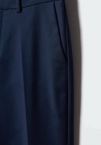 Pantalon bleu marine foncé sur mesure suspendu sur un fond clair, montrant la ceinture avec passants pour ceinture et détail de la poche latérale.