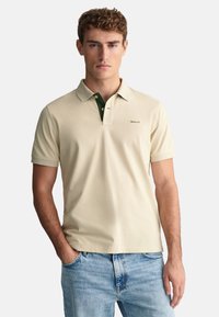 Beige polo skjorta med grön krage, kort ärm och slim fit. Har en liten logotyp på vänster bröst och är gjord av mjukt bomullsmaterial.