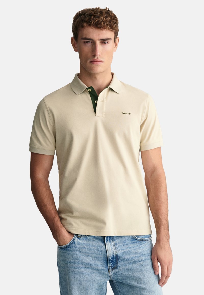 Beige polo skjorta med grön krage, kort ärm och slim fit. Har en liten logotyp på vänster bröst och är gjord av mjukt bomullsmaterial.